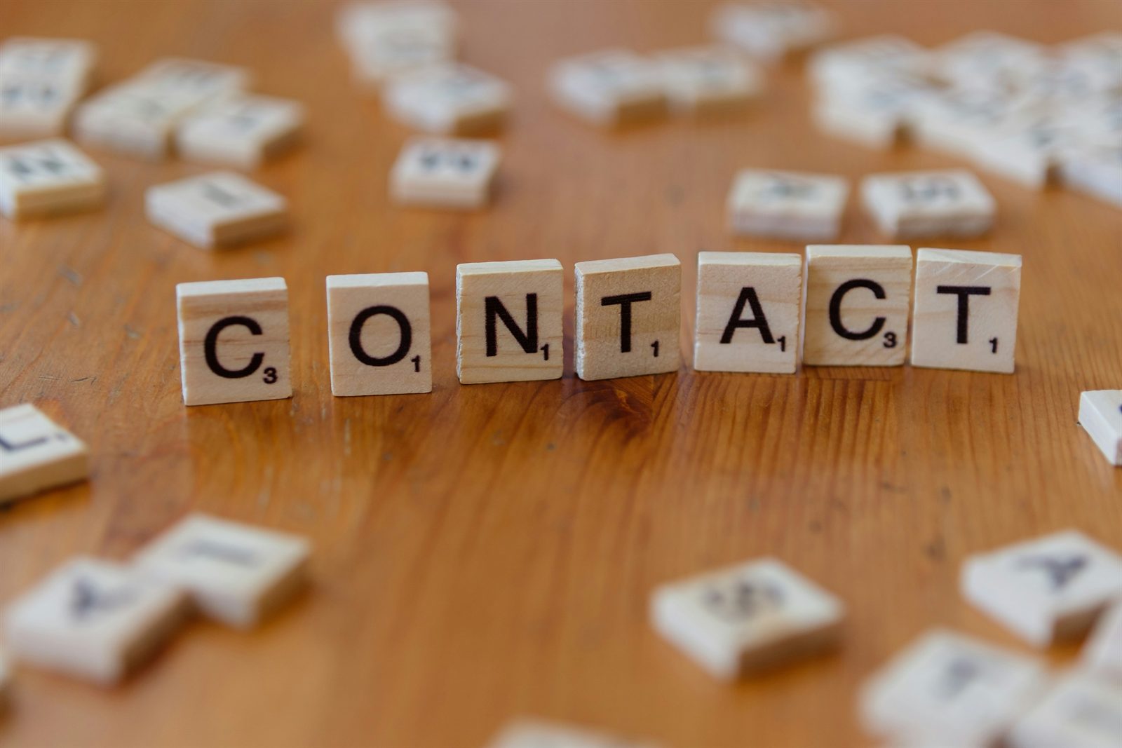 Scrabble letter tiles arranged on a wooden table to spell the word CONTACT.
