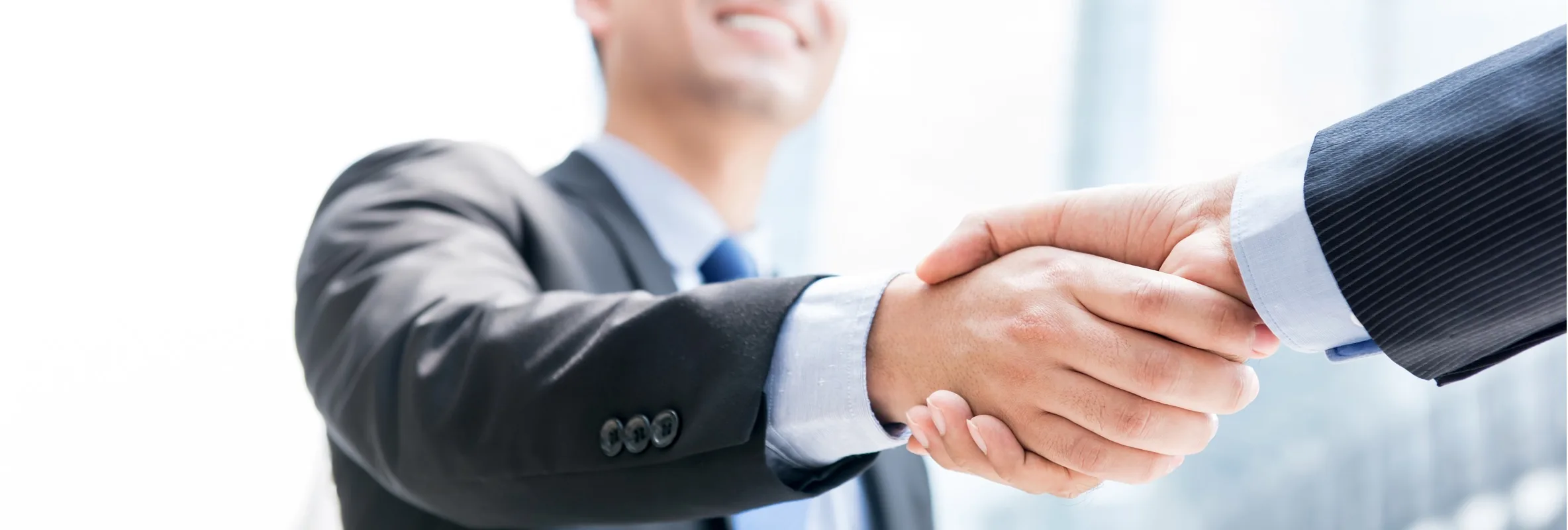 Two people in business suits shaking hands in front of a bright window.